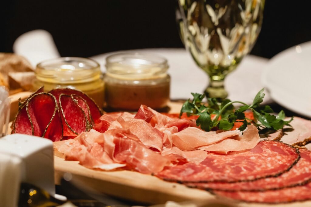 Closeup of delicious sliced bacon meat and salami served on table with wineglass and mustard on blurred background of white plates in dark room